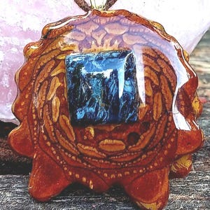 May include: A pendant featuring a brown, textured pine cone scale base coated in a glossy resin finish. A square, deep blue, chatoyant gemstone is embedded in the center. The pendant hangs from a brown cord, creating a circular jewelry piece.