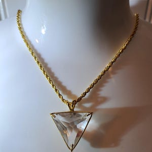 Op de afbeelding: Een goudkleurige ketting met een heldere, driehoekige hanger. De hanger is facetgeslepen en vangt het licht. De ketting heeft een gedraaid touwontwerp. De ketting wordt op een witte ondergrond getoond.