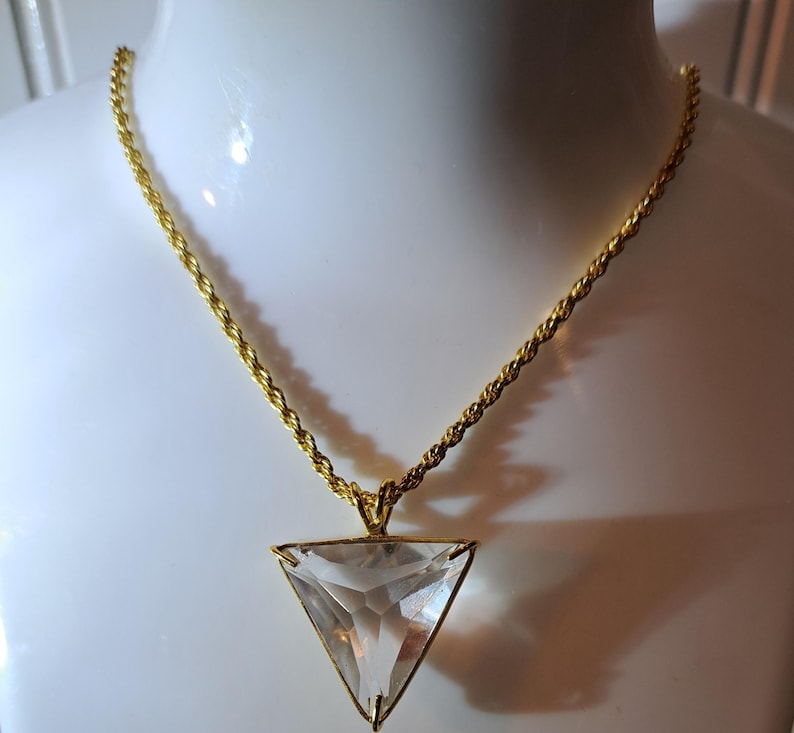 Op de afbeelding: Een goudkleurige ketting met een heldere, driehoekige hanger. De hanger is facetgeslepen en vangt het licht. De ketting heeft een gedraaid touwontwerp. De ketting wordt op een witte ondergrond getoond.