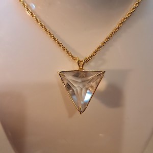 Op de afbeelding: Een goudkleurige ketting met een heldere, driehoekige hanger. De hanger is gevat in een gouden frame en hangt aan een gouden touwketting. De facetten van de hanger vangen het licht op, waardoor een sprankelend effect ontstaat. De ketting wordt gepresenteerd tegen een neutrale achtergrond.