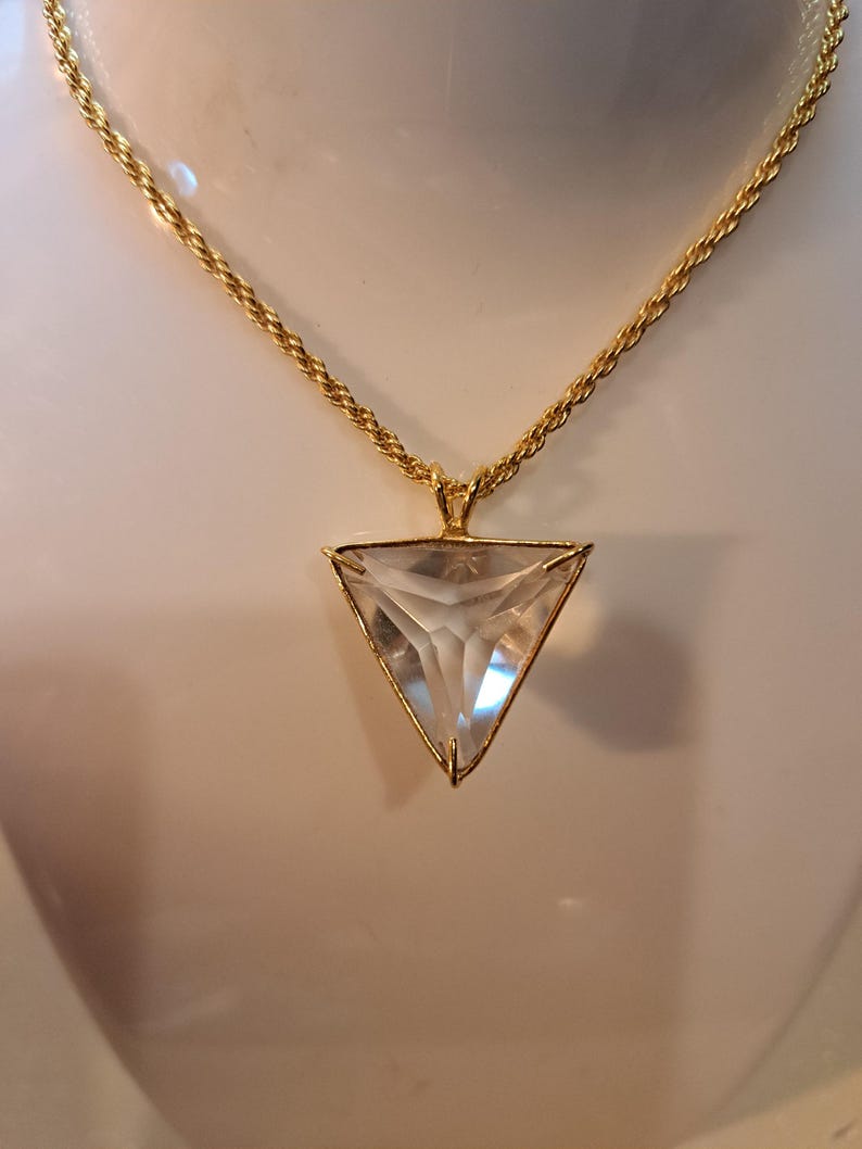 Op de afbeelding: Een goudkleurige ketting met een heldere, driehoekige hanger. De hanger is gevat in een gouden frame en hangt aan een gouden touwketting. De facetten van de hanger vangen het licht op, waardoor een sprankelend effect ontstaat. De ketting wordt gepresenteerd tegen een neutrale achtergrond.
