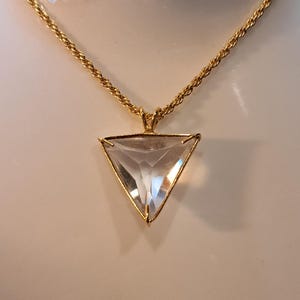 Op de afbeelding: Een goudkleurige ketting met een heldere, driehoekige hanger. De hanger is facetgeslepen en vangt het licht. De ketting heeft een gedraaid touwontwerp. De ketting wordt gepresenteerd op een wit oppervlak.
