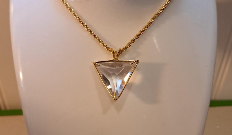 Op de afbeelding: Een goudkleurige ketting met een heldere, driehoekige hanger. De hanger is facetgeslepen en vangt het licht. De ketting heeft een gedraaid touwontwerp. De ketting wordt gepresenteerd op een wit oppervlak.