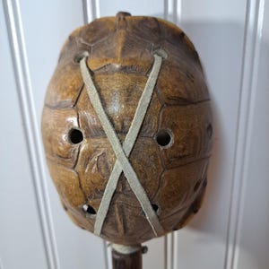May include: A vintage turtle shell mask with a dark brown wooden handle. The mask is a light brown color with a criss-cross light green strap. The mask has several holes and a textured surface.