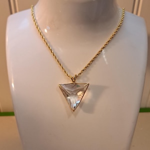 Op de afbeelding: Een goudkleurige ketting met een heldere, driehoekige hanger. De hanger is facetgeslepen en vangt het licht op. De ketting wordt getoond op een witte mannequin buste, die het ontwerp van de sieraden benadrukt. De ketting heeft een gedraaid touwontwerp.