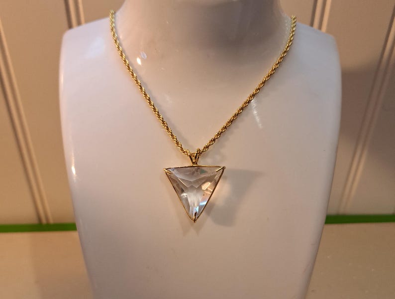 Op de afbeelding: Een goudkleurige ketting met een heldere, driehoekige hanger. De hanger is facetgeslepen en vangt het licht op. De ketting wordt getoond op een witte mannequin buste, die het ontwerp van de sieraden benadrukt. De ketting heeft een gedraaid touwontwerp.