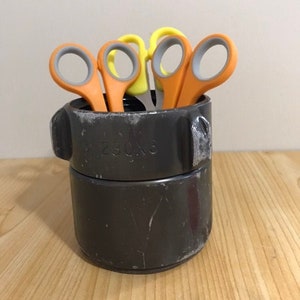 Könnte beinhalten: Ein dunkelgrauer zylindrischer Behälter mit mehreren Scherenpaaren. Die Scheren haben orange und gelbe Griffe mit grauen Fingerlöchern. Der Behälter steht auf einer Holzoberfläche.