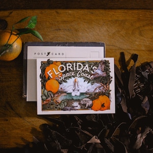 May include: A vintage postcard featuring a colourful illustration of a rocket launching from a beach with palm trees and oranges. The text on the postcard reads "Florida's Space Coast".