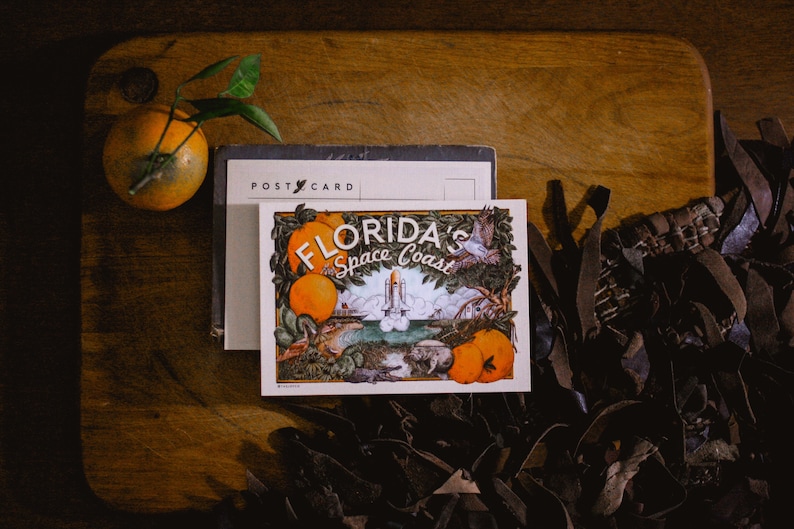 May include: A vintage postcard featuring a colourful illustration of a rocket launching from a beach with palm trees and oranges. The text on the postcard reads "Florida's Space Coast".