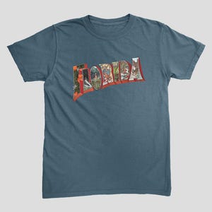May include: Teal-colored t-shirt featuring the word "FLORIDA" in a vintage postcard style. The letters are filled with images of Florida landscapes and landmarks. The shirt has short sleeves and a crew neck.