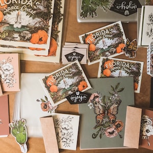 May include: A flat lay of various items including cards, stickers, and prints. The cards feature floral designs and the text "FLORIDA'S Space Coast". The stickers feature butterflies, flowers, and cacti. The prints feature a butterfly with flowers and the text "the joy.co."