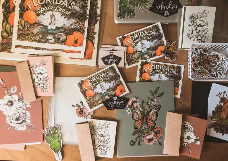 May include: A flat lay of various items including cards, stickers, and prints. The cards feature floral designs and the text "FLORIDA'S Space Coast". The stickers feature butterflies, flowers, and cacti. The prints feature a butterfly with flowers and the text "the joy.co."