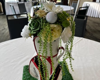 May include: A white, red, and black miniature golf bag centerpiece with the text GOGOGO printed on the side. The bag is filled with an arrangement of white and green artificial flowers, greenery, and a single white golf ball on top.