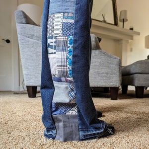May include: Close-up of the bottom of a pair of blue jeans with a patchwork design on the side seam. The patchwork is made of various fabrics in shades of blue, gray, and white.