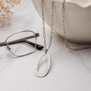 May include: A silver necklace with an oval, hammered-texture pendant. The necklace is displayed near a pair of silver-framed glasses and a white ceramic bowl. The background features dried flowers on a white marble surface.