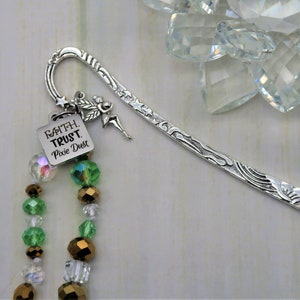 May include: Silver bookmark with a fairy charm and a beaded chain. The beads are green, gold, and clear. The charm has the words "Faith Trust Pixie Dust" on it.