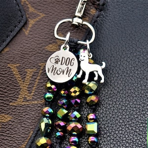 May include: A silver charm with the words "Dog Mom" and a paw print inside a heart, attached to a silver dog charm and a beaded chain with iridescent rainbow colored beads. The charm is attached to a black leather bag.