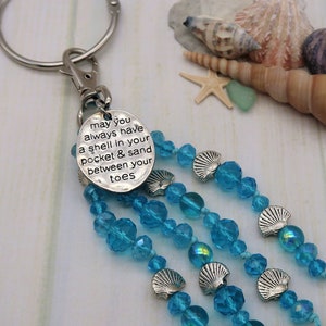Pode incluir: Um chaveiro com um charme de prata e um tassel de contas de cristal azul. O charme tem o texto "may you always have a shell in your pocket & sand between your toes".