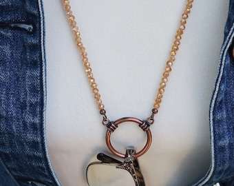 May include: A long necklace with a copper-colored pendant in the shape of an apple. The necklace has a beaded chain in a light brown color. The person is wearing a white top and a blue denim jacket. The text "Crafted by Kristal" is in the upper left corner.
