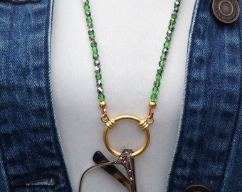 Könnte beinhalten: Eine lange Halskette mit kleinen, facettierten grünen Kristallperlen an einer Schnur, verbunden mit einem goldfarbenen Metallring zum Halten einer Brille. Die Kette wird über einem weißen Oberteil und einer blauen Jeansjacke getragen. Text: Crafted By Kristal.