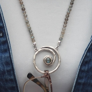 May include: A silver necklace with a spiral pendant holding eyeglasses. The necklace features a beaded chain with small, gray beads. The pendant has a small, round, blue-green stone in the center. The necklace is worn over a white top and a blue denim jacket.