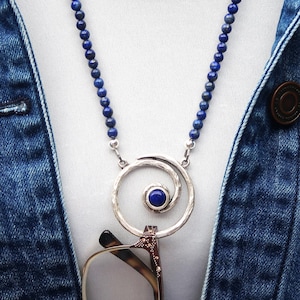 May include: A silver-toned necklace with a spiral pendant and a small, round, blue gemstone. The necklace has a beaded chain of small, dark blue beads. The necklace is holding a pair of eyeglasses. The model is wearing a denim jacket.