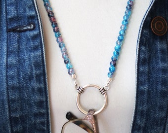 May include: A beaded eyeglass holder necklace with blue and purple beads. The necklace has a silver-colored ring to hold glasses. The necklace is worn over a white shirt and a denim jacket.