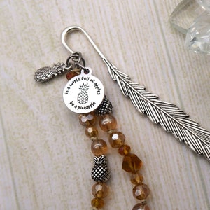 May include: Silver feather bookmark with a charm that reads "In a world full of apples be a pineapple". The charm has a pineapple graphic. The bookmark is adorned with brown glass beads and silver pineapple charms.