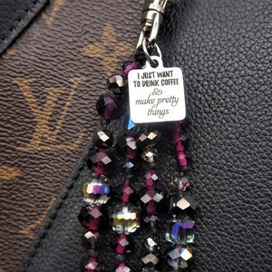 May include: A black and silver charm with the text "I just want to drink coffee & make pretty things" hanging from a beaded tassel with black, purple, and iridescent beads. The charm is attached to a brown and black patterned handbag.
