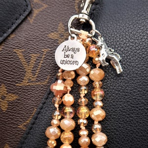 May include: A silver charm with the text "Always be a unicorn" hangs from a beaded keychain with a silver unicorn charm. The keychain is made of peach and gold beads.