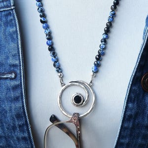 May include: A necklace with a silver pendant holding eyeglasses. The necklace features a beaded chain with blue and black beads. The pendant has a spiral design with a black stone in the center. The necklace is worn over a white shirt and a denim jacket.