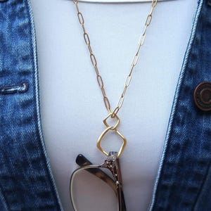 Puede incluir: Un collar dorado para sujetar gafas con un colgante geométrico. El collar sostiene un par de gafas con un patrón de carey. El fondo incluye una chaqueta vaquera y una camisa blanca.