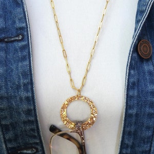 May include: A gold-colored necklace with a textured circular pendant and a long chain. The necklace is worn over a white top and a blue denim jacket. The image also shows a pair of eyeglasses.