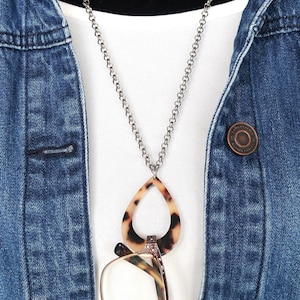 May include: A silver chain necklace with a tortoiseshell-patterned pendant that holds glasses. The pendant is shaped like a teardrop. The necklace is worn over a white shirt and a denim jacket.