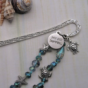 May include: Silver bookmark with a charm that says "Just Keep Swimming" and a beaded chain with sea turtle and starfish charms. The bookmark is on a white wooden surface with seashells.