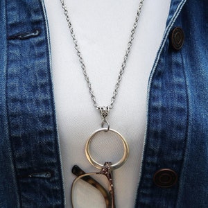 May include: A silver chain necklace with a round metal ring attached to the end. The ring is designed to hold glasses. The necklace is worn over a white shirt and a blue denim jacket.