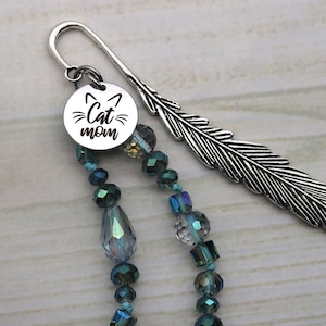 May include: A silver feather bookmark with a silver charm that says "Cat Mom" and a cat silhouette. The charm is attached to a strand of teal and clear crystal beads.
