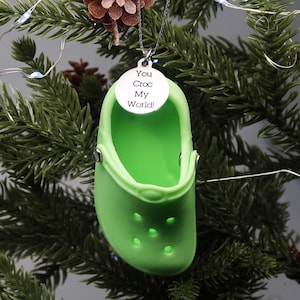 Puede incluir: Adorno navideño de plástico verde con forma de zueco con un dije plateado que dice "You Croc My World!"