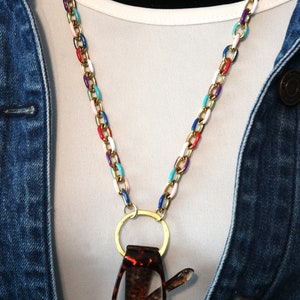 May include: A gold chain with colorful enamel links, worn as a glasses chain. The chain has a gold ring at the bottom to hold glasses. The chain is worn over a white shirt and a denim jacket.