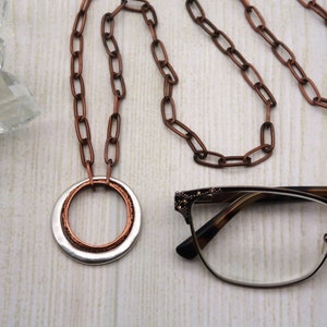 May include: A copper chain necklace with a silver and copper ring pendant. The necklace is laying on a white wooden surface with a pair of eyeglasses next to it. The text "CRAFTED BY KRISTAL" is visible in the lower left corner of the image.