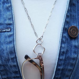 Puede incluir: Un collar plateado para sujetar gafas con un colgante geométrico. El collar se lleva sobre una camiseta blanca y una chaqueta vaquera azul. Las gafas son marrones y doradas.