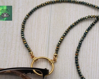 Könnte beinhalten: Brillenkette mit kleinen, irisierenden Perlen in Grün- und Goldtönen. Die Kette ist an einem goldfarbenen Ring befestigt, der zum Halten von Brillen dient. Die Brille ist schwarz und braun. Das Logo "Crafted by Kristal" befindet sich oben links.