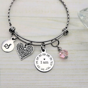 May include: A silver charm bracelet with a heart charm, a round charm with the words "I am" and a pink crystal charm. The bracelet also has a small round charm with the letter "S" on it.