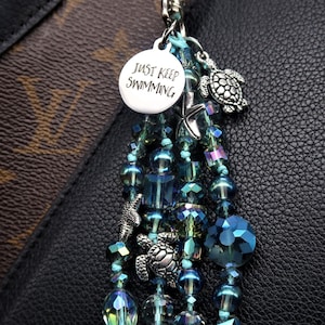 May include: A teal blue and silver charm with a silver charm that says "Just Keep Swimming" and a silver turtle charm. The charm is attached to a beaded chain with teal blue, green, and silver beads.