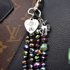 May include: A silver charm with a heart-shaped tag that reads "be a unicorn in a field of horses" and a small unicorn charm hangs from a beaded keychain with iridescent purple, green, and gold beads. The keychain is attached to a black leather bag.