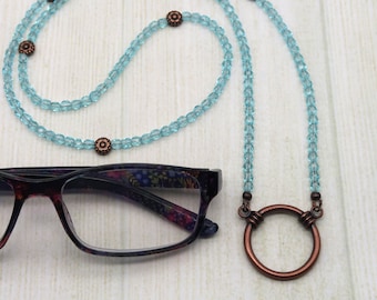 May include: A light blue beaded eyeglass chain with copper accents and a copper circle at the end. The chain is laying on a wooden surface next to a pair of eyeglasses with a floral pattern.