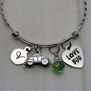 May include: A silver bangle bracelet with charms, including a car charm, a heart charm with the text "LOVE BUG", a green crystal bead, and a round charm with the letter "L".