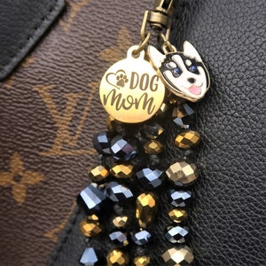 May include: Gold charm with a black and white husky dog face and the words "Dog Mom" with a paw print and heart. The charm is attached to a black and gold beaded tassel.