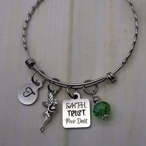 Könnte beinhalten: Silbernes Armband mit einem silbernen Anhänger, auf dem "Faith, Trust, Pixie Dust" steht, und einem grünen Kristallanhänger. Das Armband hat außerdem einen silbernen Anhänger mit dem Buchstaben "L" und einen silbernen Feenanhänger.