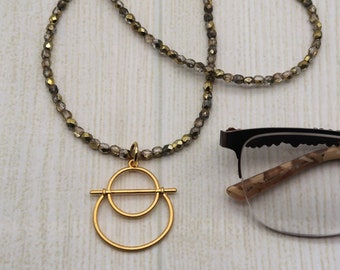May include: A beaded necklace with a gold pendant featuring two connected circles and a horizontal line. The necklace is composed of small, faceted beads in shades of brown and gold. A pair of eyeglasses rests on the right side of the image.
